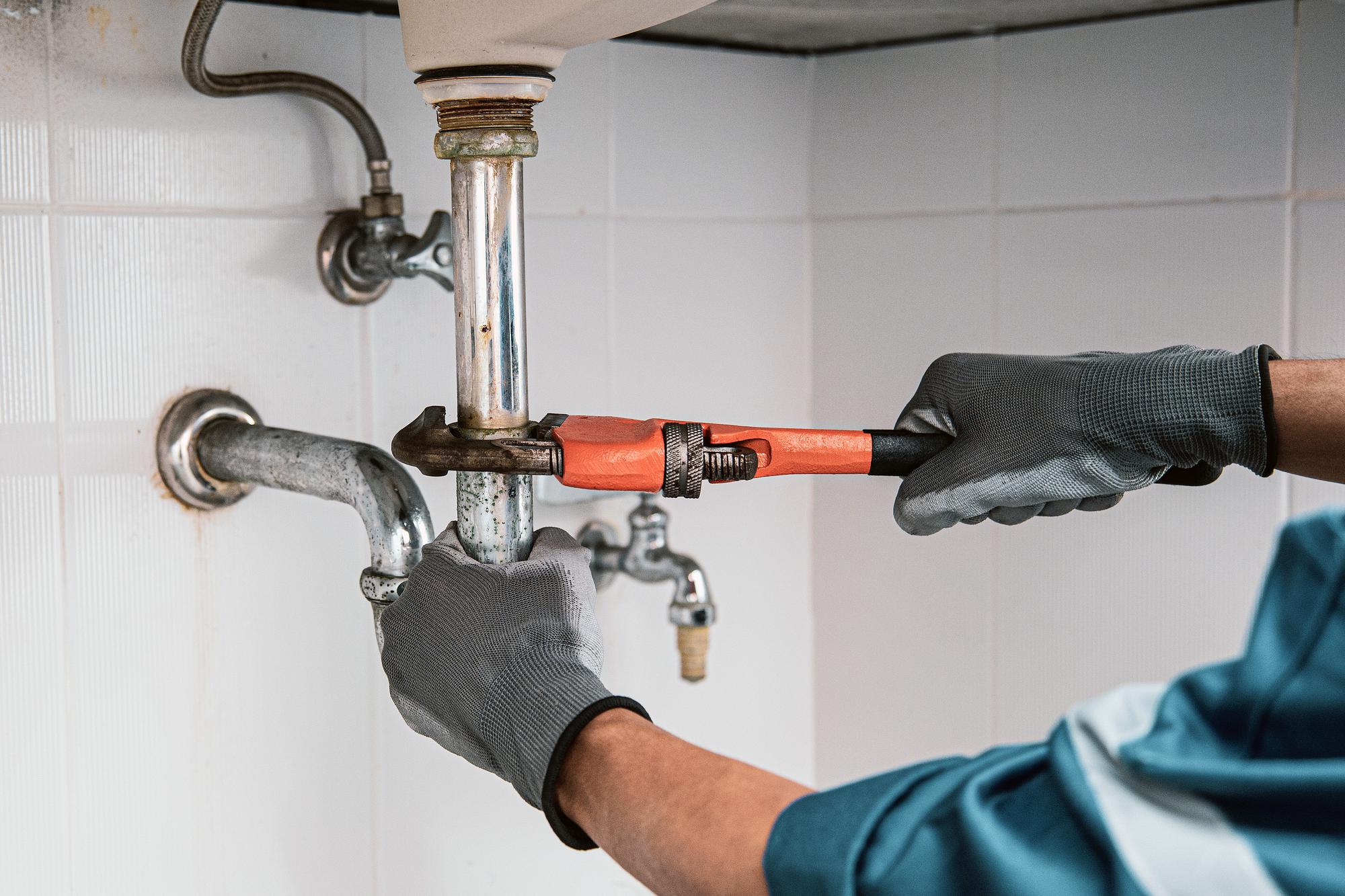 Plumber in gloves using a pipe wrench to tighten a corroded sink drain pipe in a tiled bathroom setting.