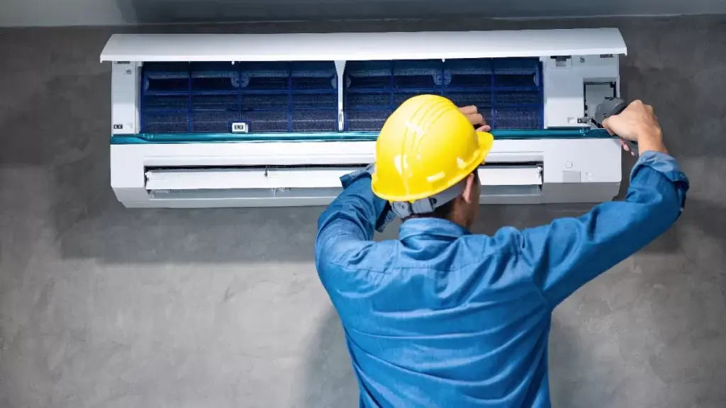 Technician in blue uniform and yellow helmet servicing a split air conditioner mounted on a grey wall.