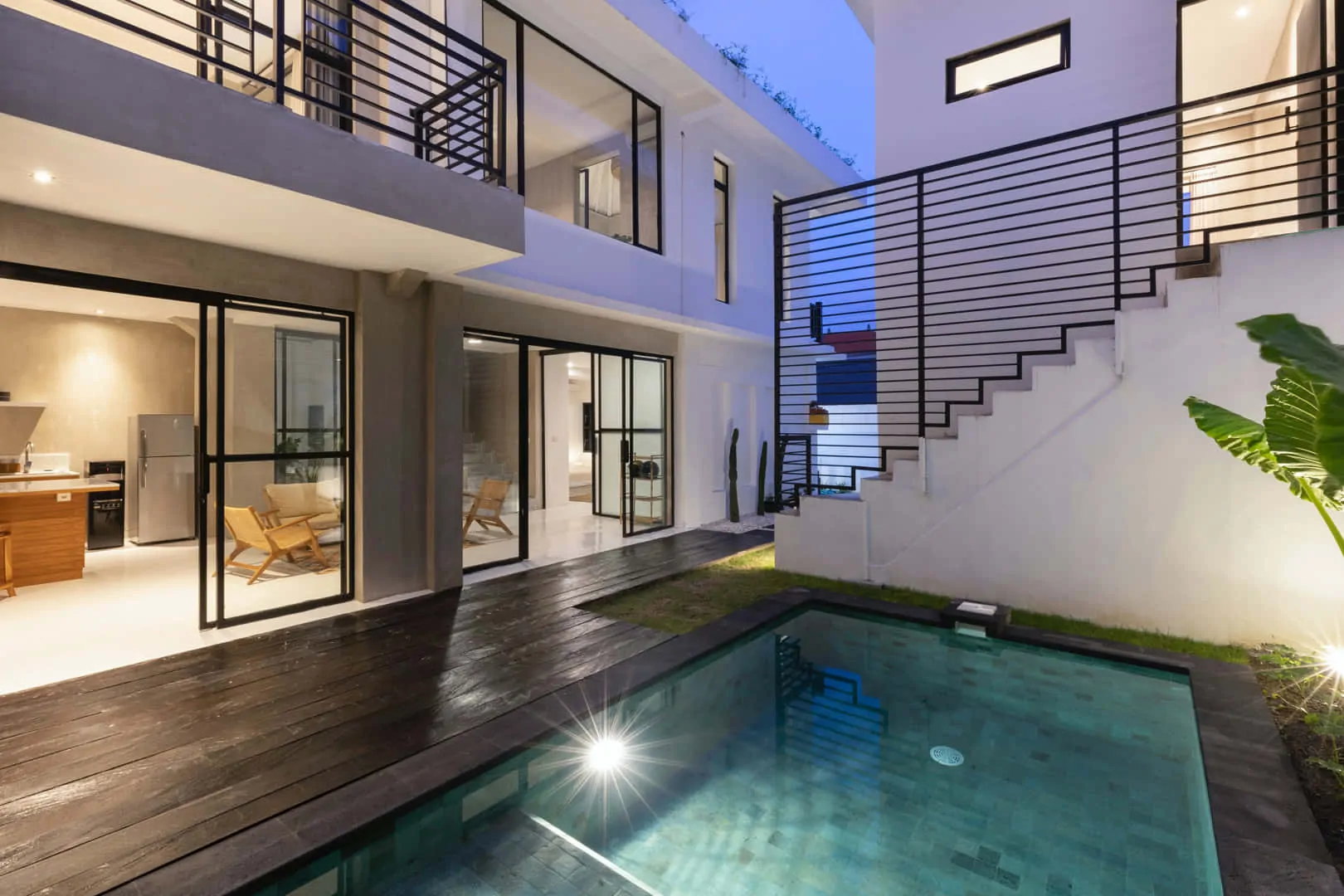 Modern villa with glass doors, wooden deck, and lit swimming pool at dusk, featuring sleek black railings.