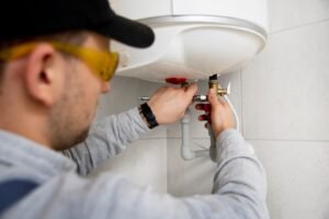 Technician in safety glasses and cap using tools to repair or install a wall-mounted water heater system.